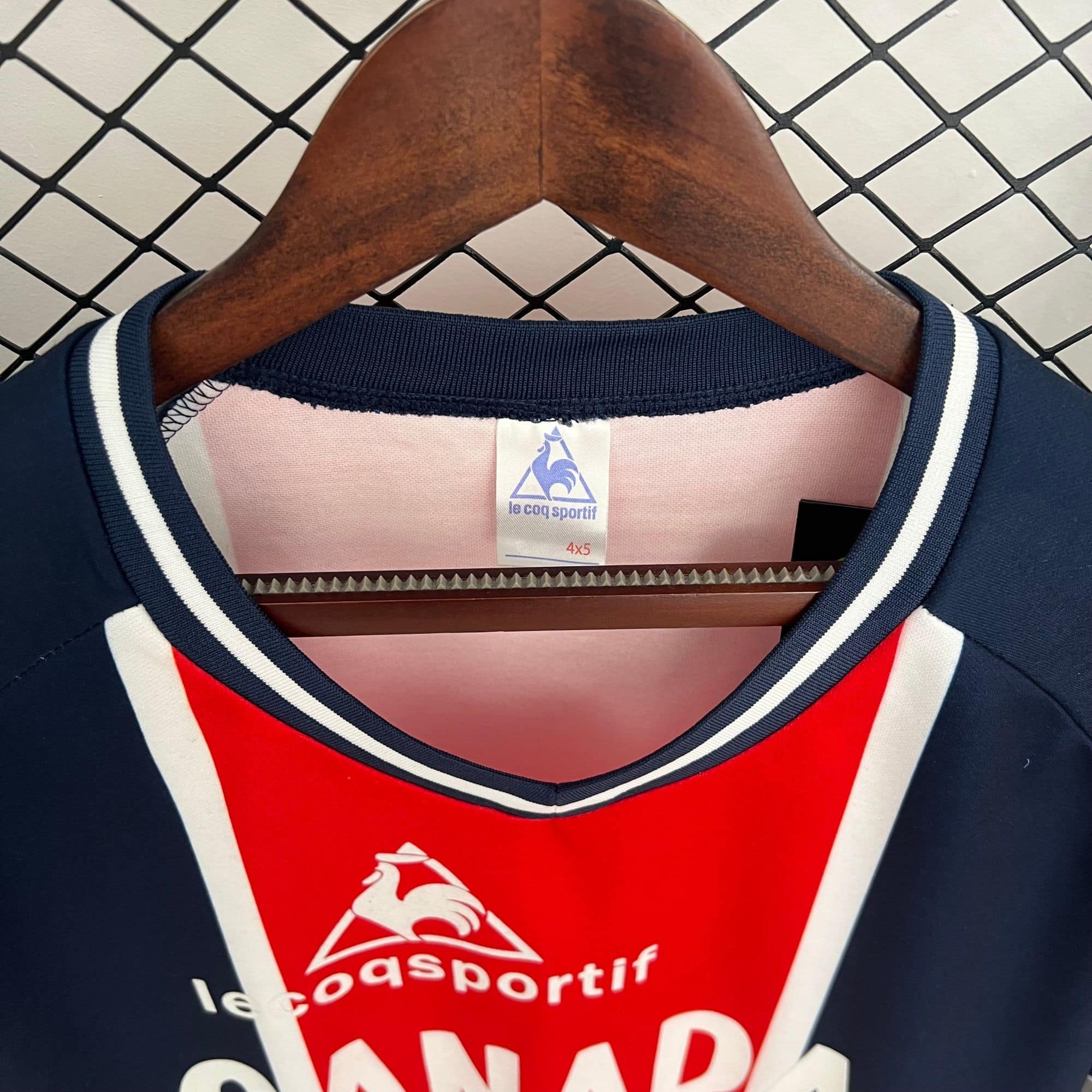 48e9802c_46e41cf5-061a-4776-a56c-b07064a6306d | AE JERSEY Close-up of a wooden hanger holding a pink Le Coq Sportif tag with the brand rooster logo and size 4x5.