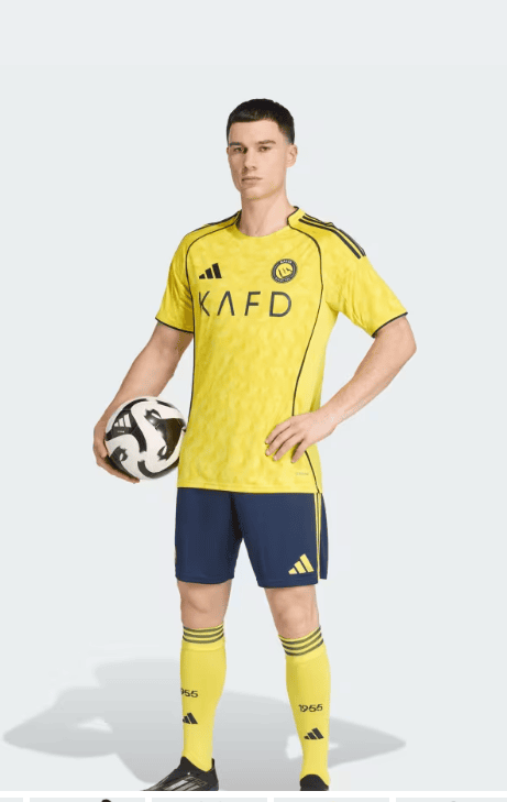 4A | AE JERSEY Full-body shot of a male soccer player in a yellow jersey and blue shorts, standing with a soccer ball under his left arm.