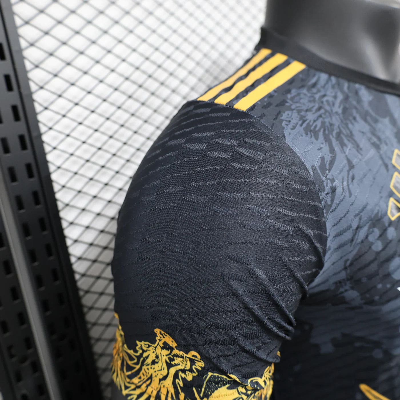 4a8caf32 | AE JERSEY Close-up of a black athletic jersey sleeve with a yellow dragon graphic and three yellow shoulder stripes against a metal grid background.