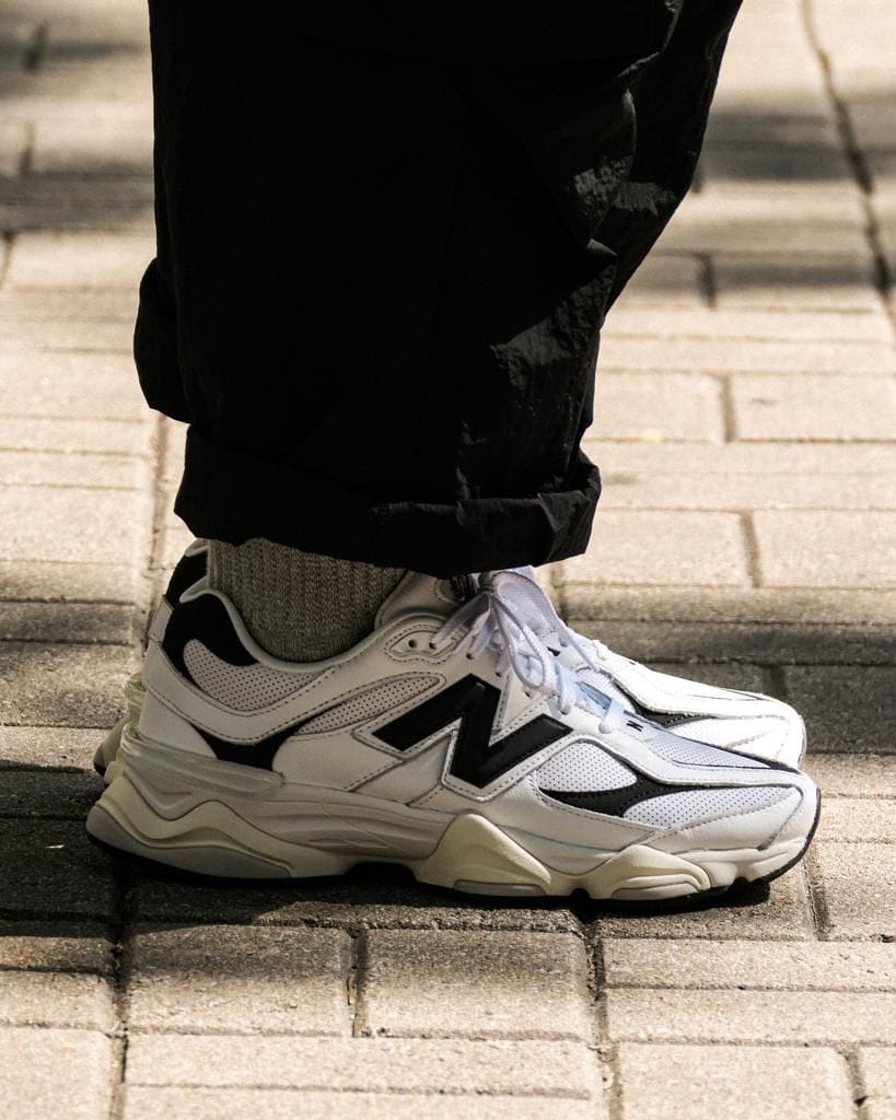 4b2a7607-4c47-4301-8f78-f8c28abb446f_b0b64d91-bf3e-4526-bd70-4be6d26e83ca | AE JERSEY White athletic sneaker with black detailing on a gray sock, worn with rolled black pants on a paved sidewalk.