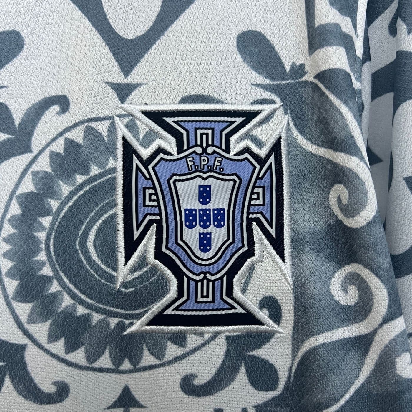 4b5d7589-8af7-45da-b633-d7a1283b959f | AE JERSEY Embroidered patch of the Portuguese Football Federation (FPF) crest on a blue and white patterned fabric background.