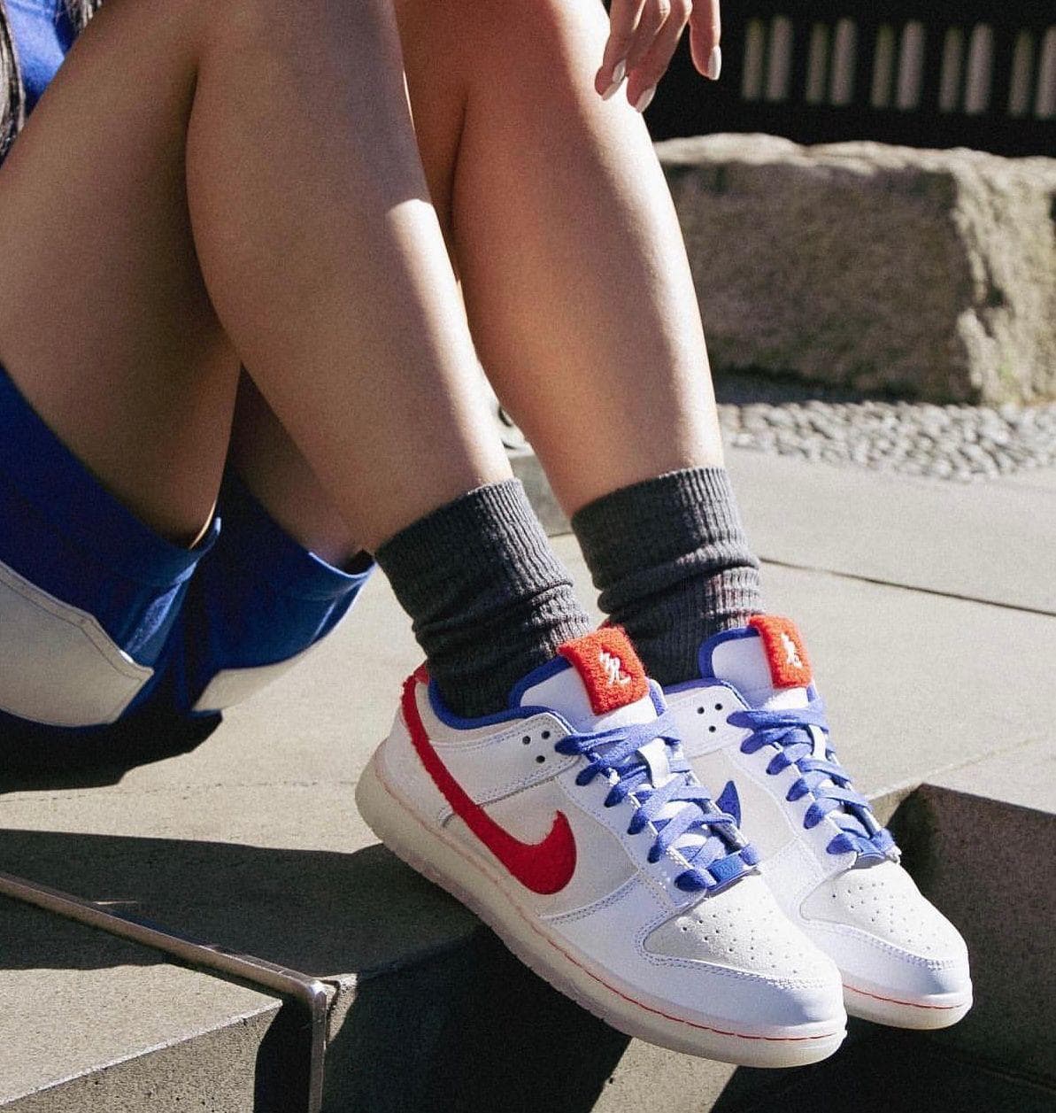 4dbbccb9-5ff6-4ea8-a641-b324ee730b1e_3cad527c-515b-4b92-a806-2a7701b94fbe | AE JERSEY Person wearing white Nike sneakers with blue laces and an orange tongue, sitting on concrete steps with gray socks showing.