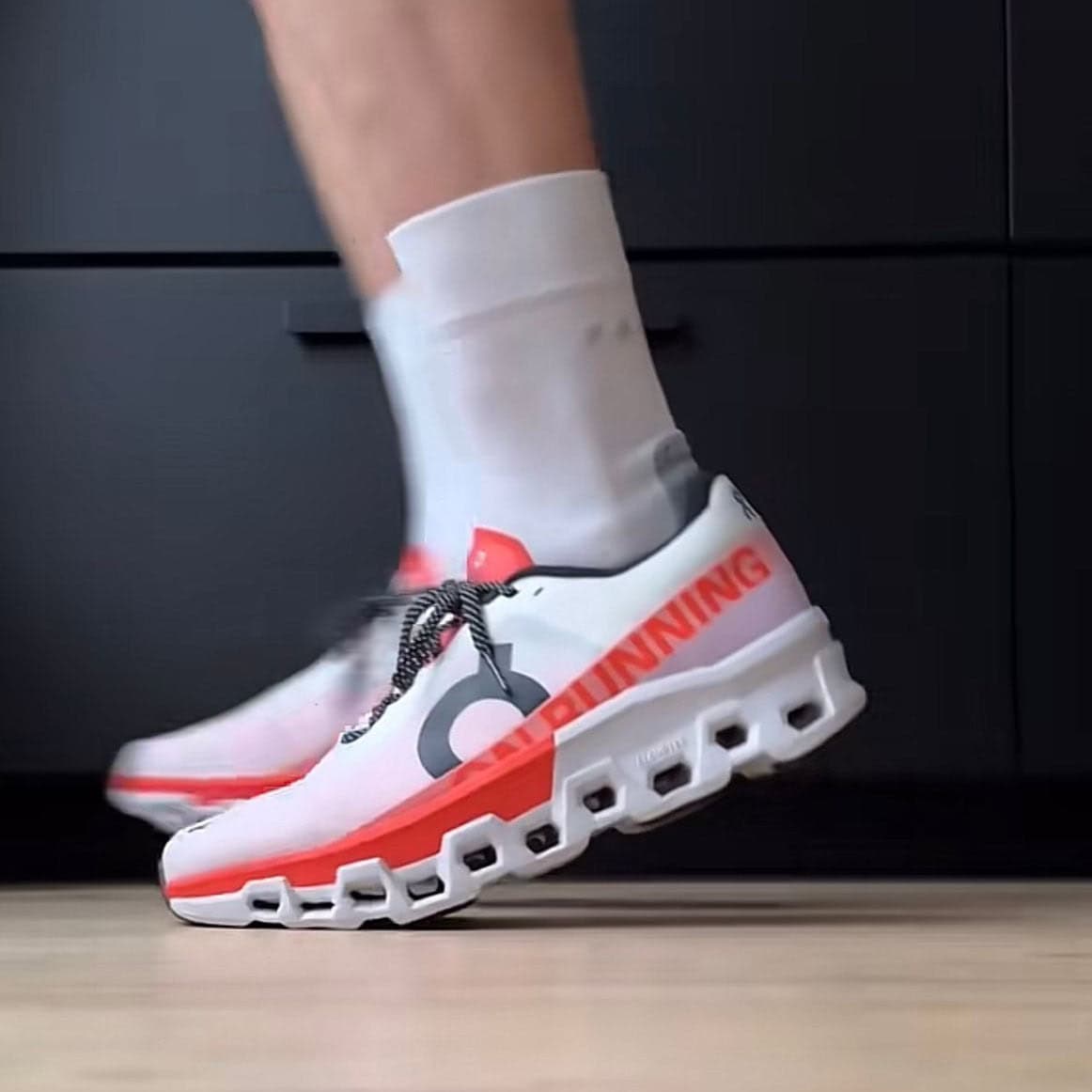 4e94c0d2-a13b-4051-967b-84f6779f19a7 | AE JERSEY White running shoe with orange accents and a chunky segmented sole mid-stride on a wooden floor, socked ankle visible in the background.