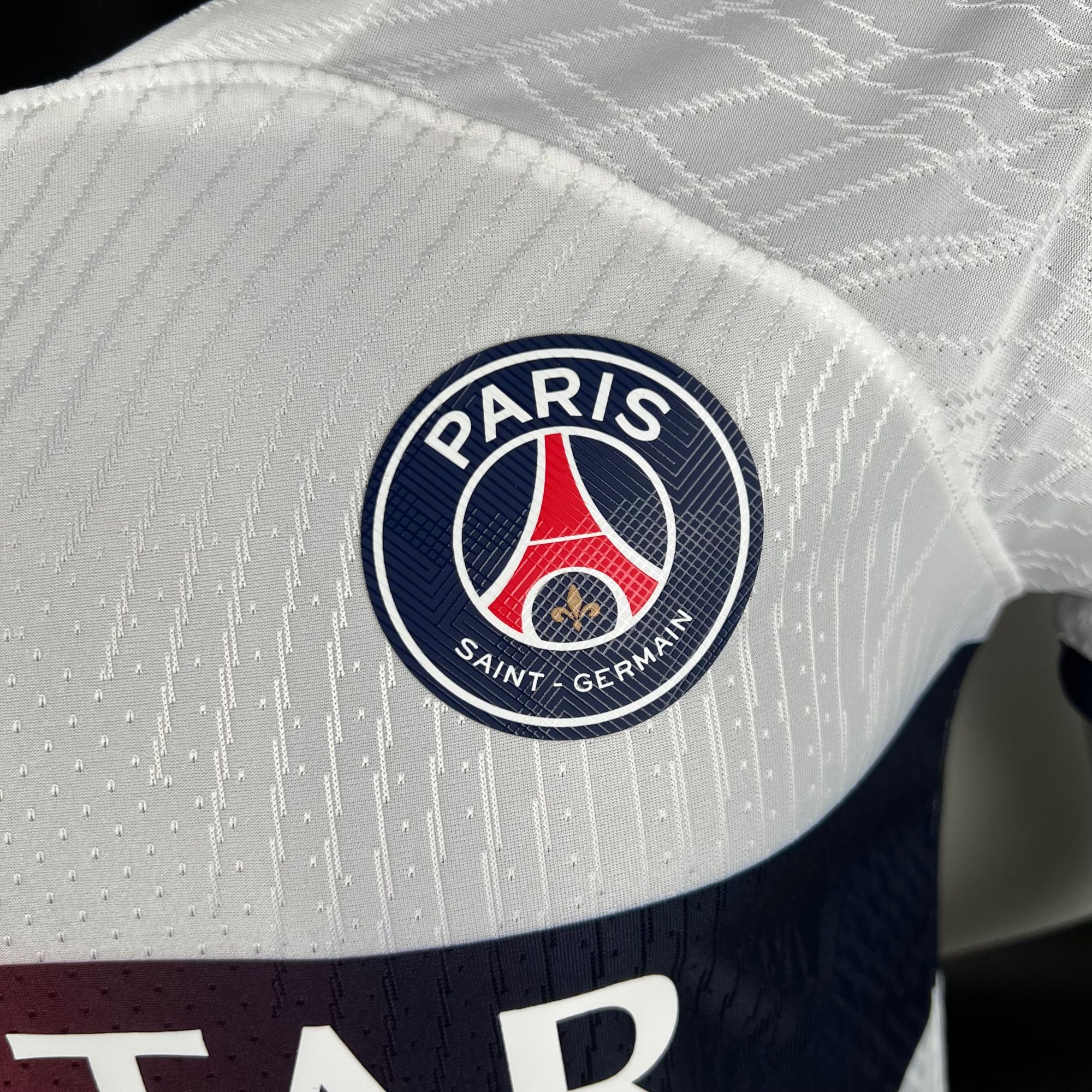 4fb5789a_a8f14b91-693d-4a29-bd79-d228fb3953a4 | AE JERSEY Close-up of the Paris Saint-Germain crest on quilted white fabric with a geometric pattern.