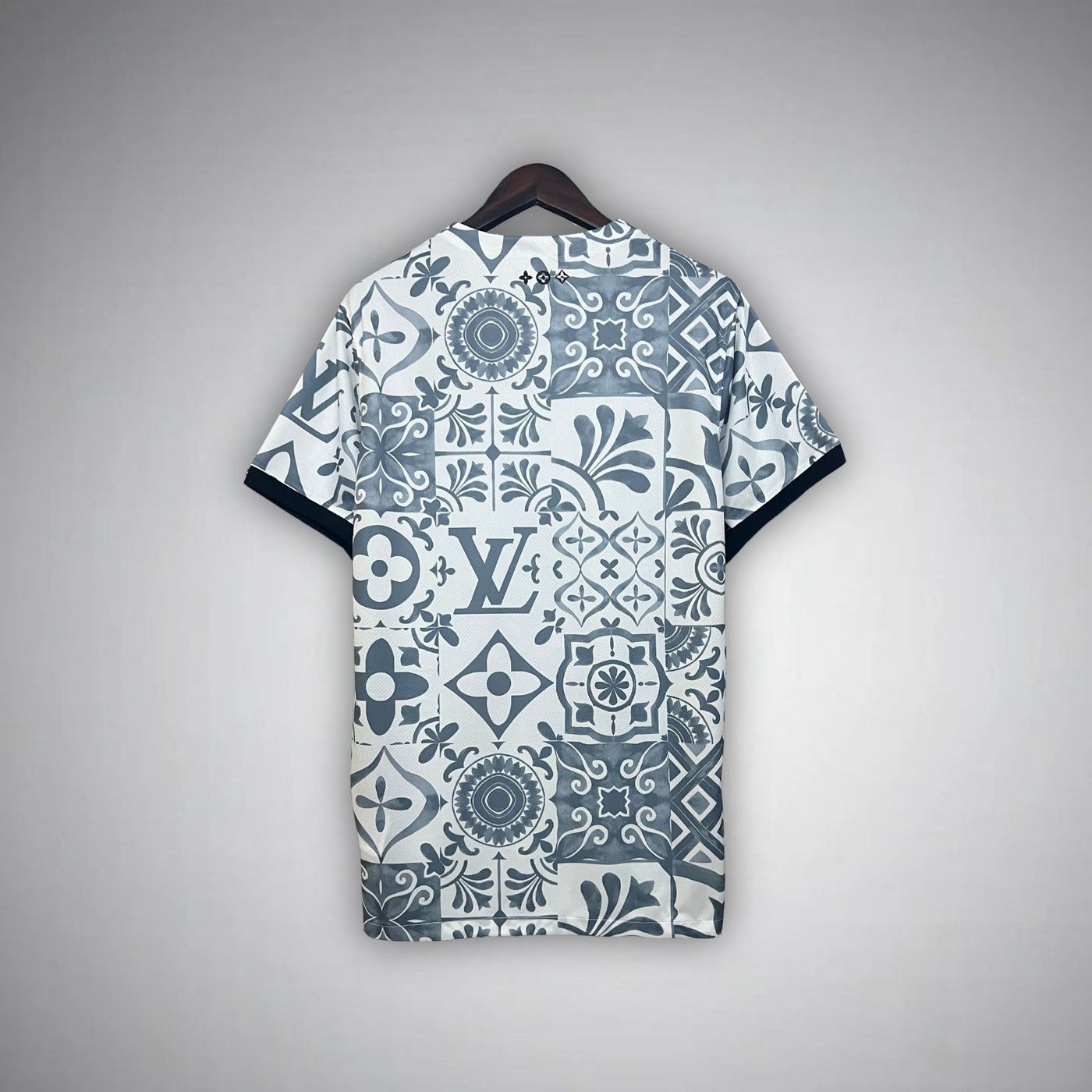 5052a1fb-0548-4d0c-94c9-d7934f5eb9bd | AE JERSEY Back view of a short-sleeve shirt on a wooden hanger, featuring a blue-gray patchwork tile pattern with floral motifs and a prominent LV monogram.