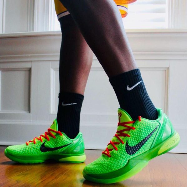 Bright neon green Nike basketball shoes with red laces, worn with black Nike socks, standing on a wooden floor by a window.