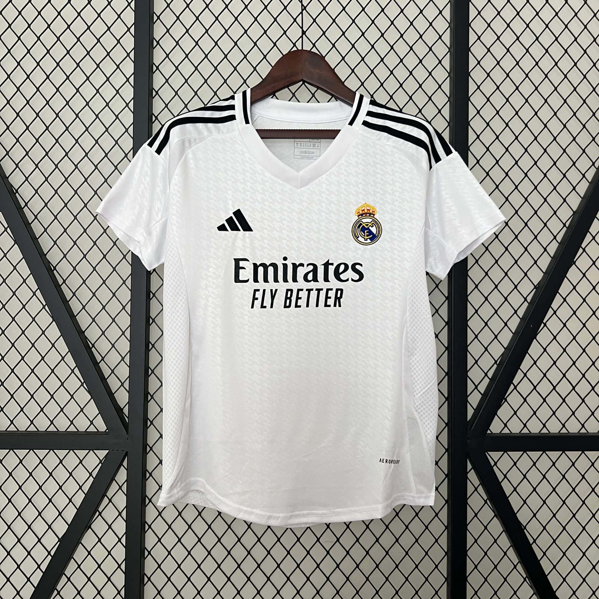 5376a8de | AE JERSEY White Real Madrid jersey on a hanger against a diamond-pattern metal grid, showing the crest, Adidas logo, and Emirates sponsor text.