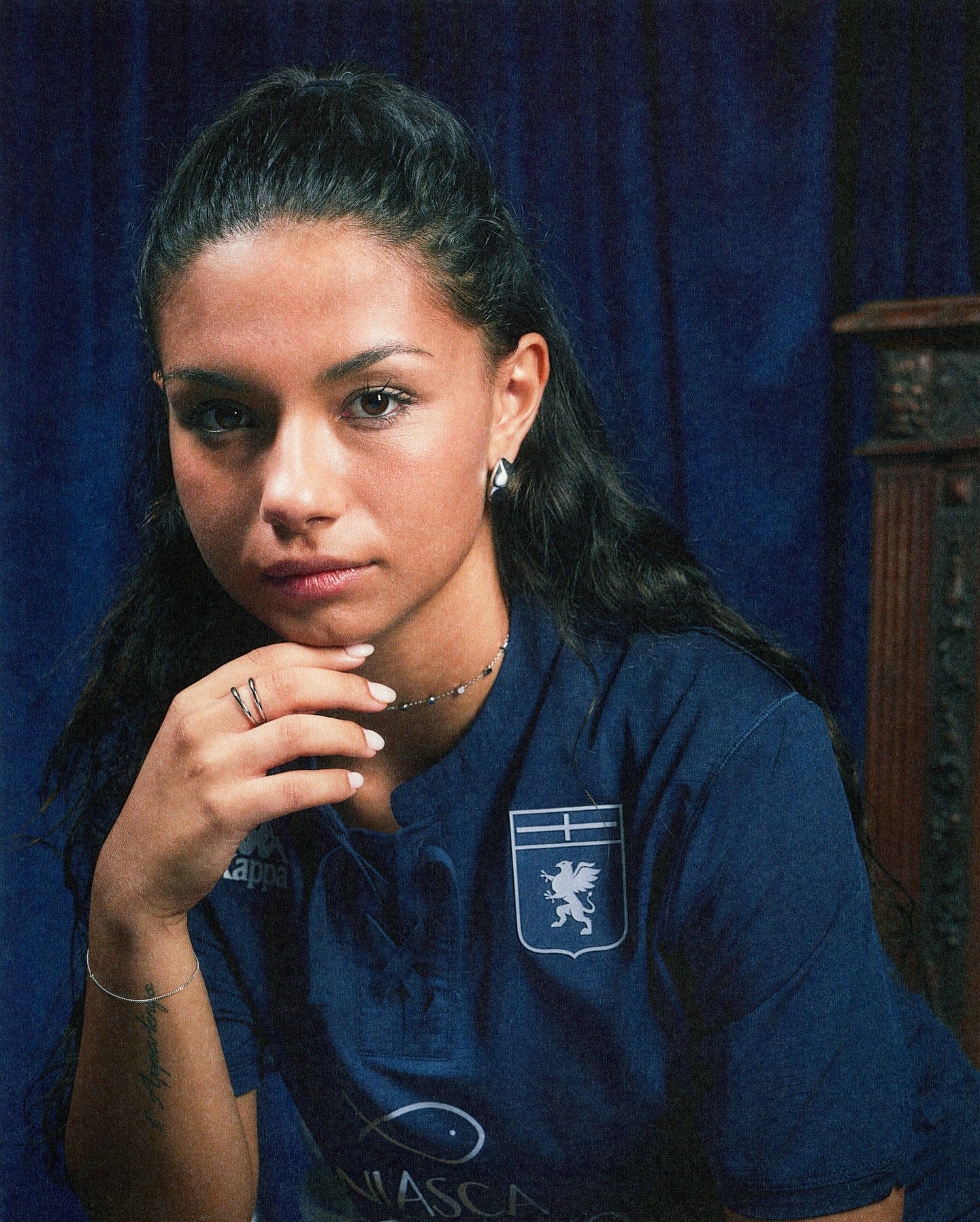 543422499_18528305539053657_1820969598630553626_n | AE JERSEY Portrait of a young woman in a blue uniform with a crest, resting her chin on clasped hands against a dark blue curtain backdrop, looking at the camera.