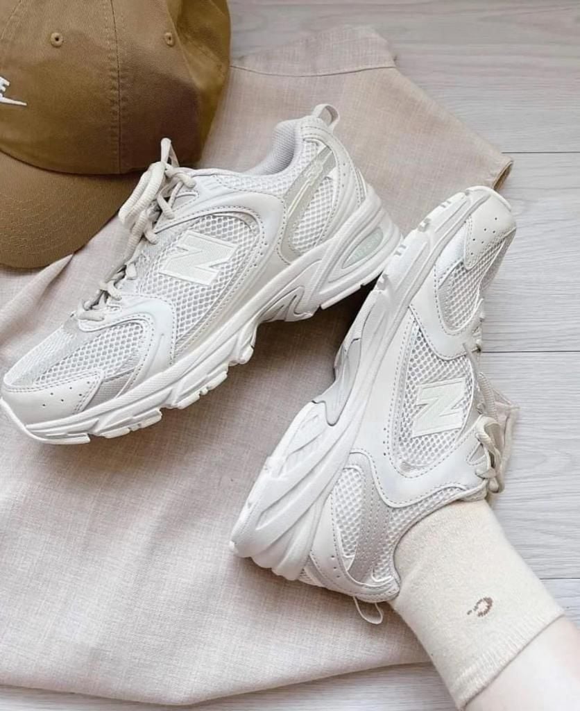 56d278a2-11ac-4ae1-8728-5017ecea7821 | AE JERSEY White chunky athletic sneakers with mesh panels, resting on light beige fabric with a brown cap nearby.