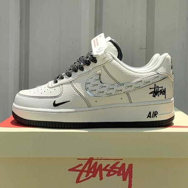 White Nike Air Force 1 sneaker with black Swoosh, Stussy logo details on the side, and a patterned strap-like overlay; shown on a boxed display.