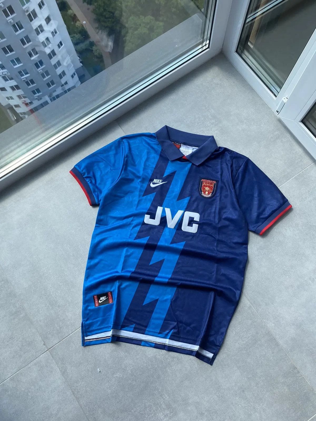 598f3429128643e3b89d5387bf44b182 | AE JERSEY Blue soccer jersey laid flat on a light gray tiled floor beside a glass door, featuring a zigzag lighter blue pattern, Nike logo, JVC sponsor, and team crest on the chest.