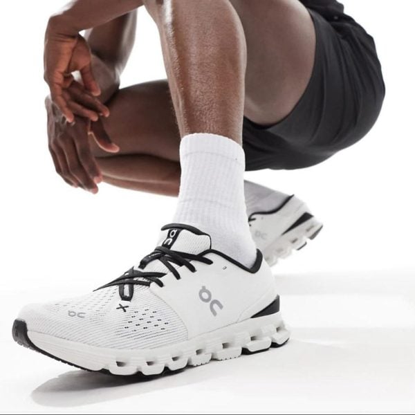 Close-up of a white running shoe with black laces and a chunky sole on a person wearing white socks.