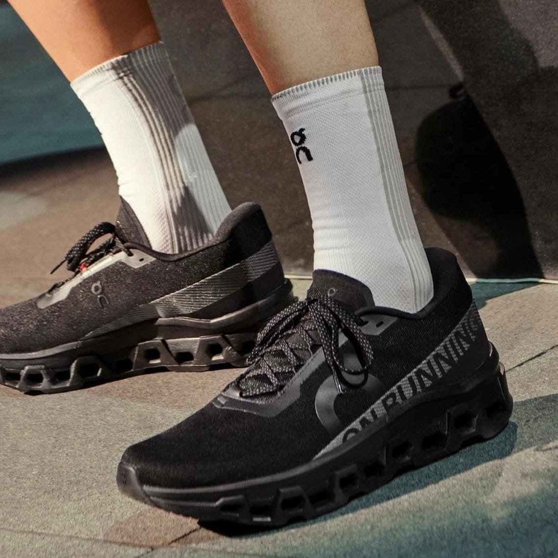5dc66427-ac66-4bd9-a6a5-94aa16edb86f | AE JERSEY Black On running sneakers with a sculpted outsole, paired with white socks, standing on a paved surface.
