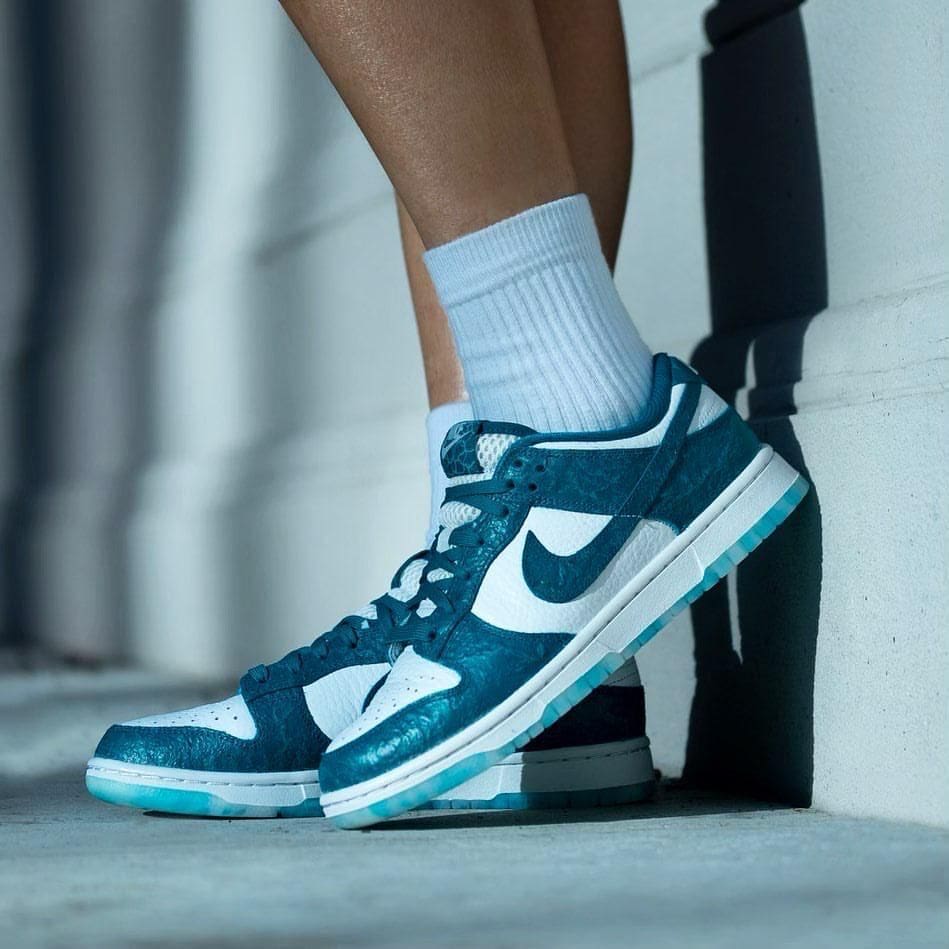 5fd9e2f6-e481-400f-9dc0-9e523adeb0df_5cce535d-3a34-4efc-b279-19344a2d0859 | AE JERSEY Teal and white Nike sneaker on a white sock, resting against a concrete wall.