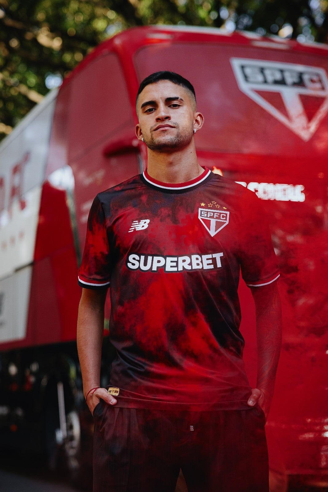 5tThBogOclh4LfK | AE JERSEY Man in a red and black São Paulo FC jersey with SUPERBET sponsor, standing with hands in pockets in front of a red team's van.