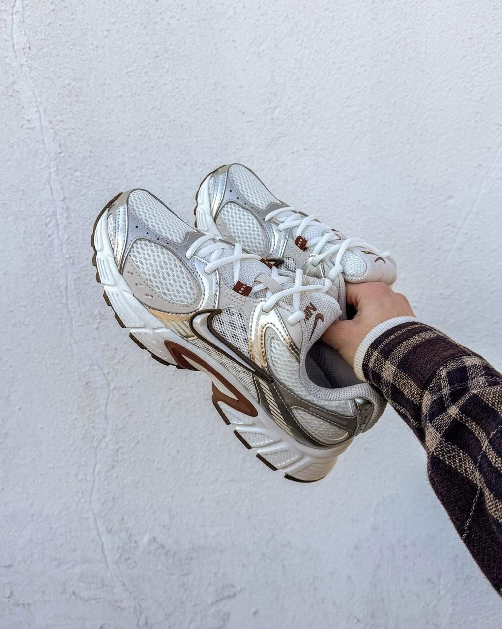 60537eff-5438-4b49-a16b-f81eba7f380e | AE JERSEY Person in a plaid sleeve holds a pair of white and gray running shoes against a pale wall.