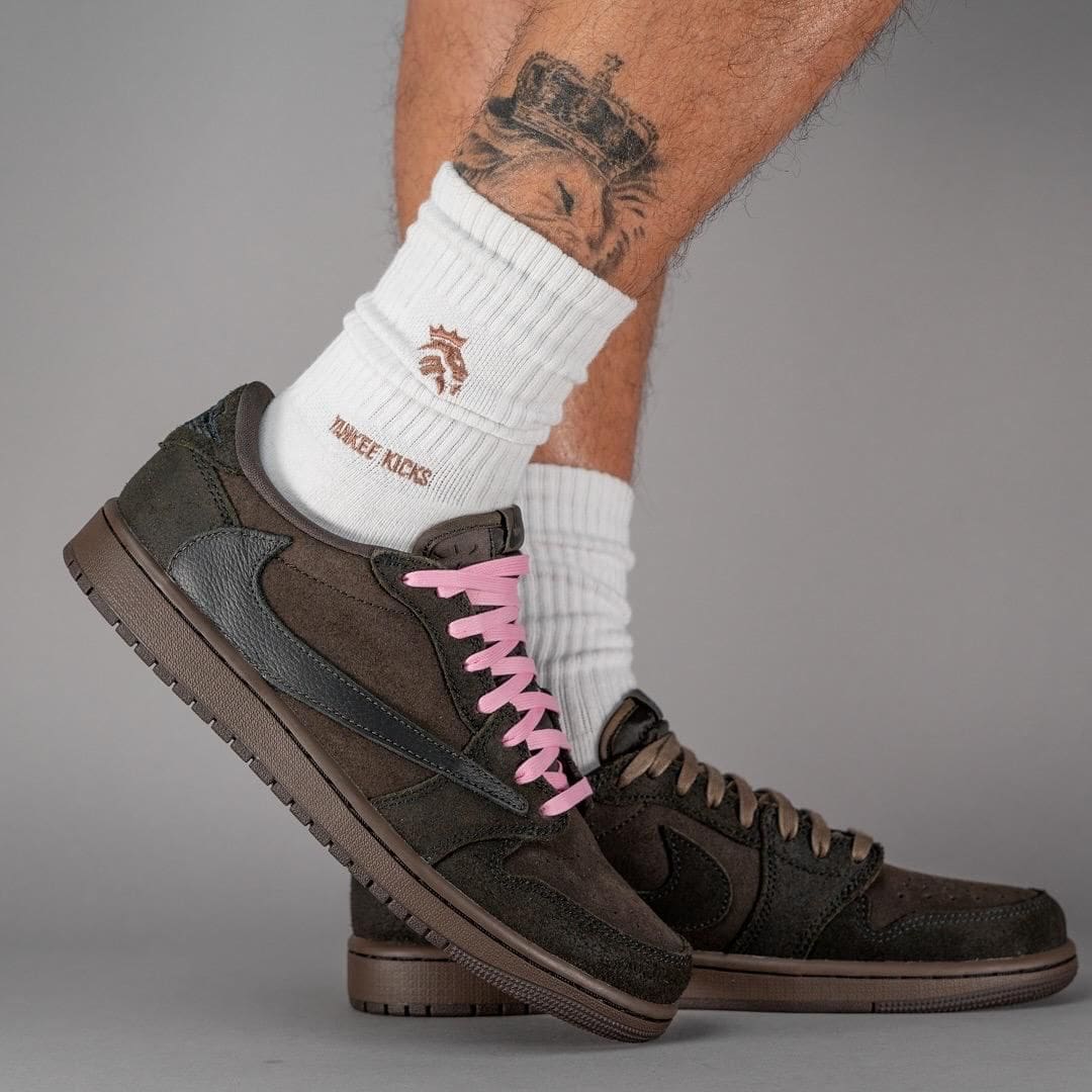 60eff74a-45c4-45c0-b7d7-cafe7d81d30e | AE JERSEY Person wearing brown suede Nike sneakers with pink laces and white crew socks, shown from the side with a tattooed leg in the background.