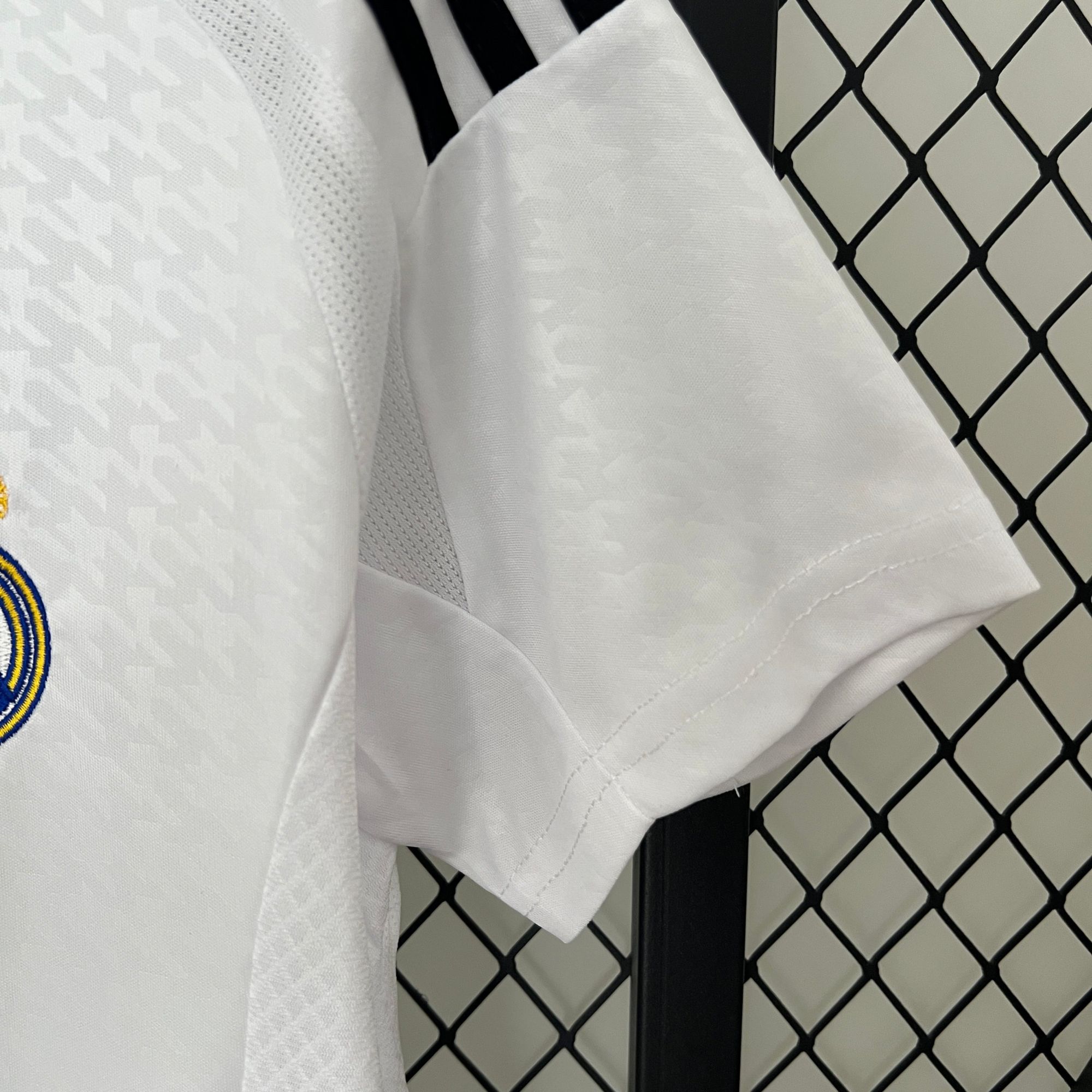 62b2fedc | AE JERSEY Close-up of white fabric with stitching, a gray seam running down the center, and a black metal post with a chain-link fence in the background.