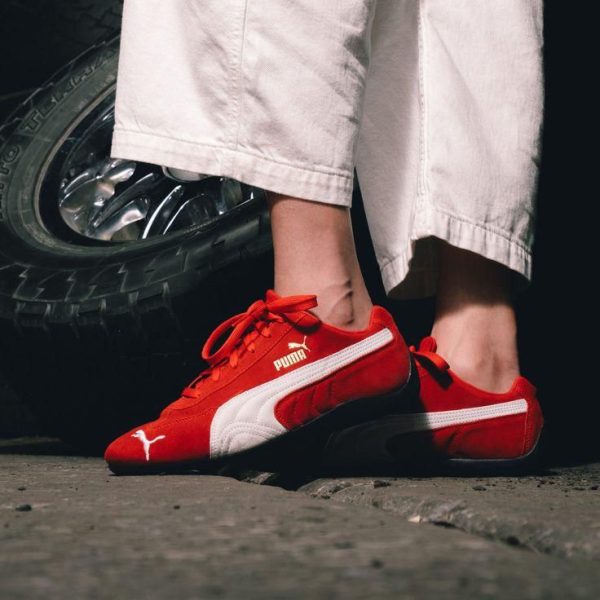 62f9a22d-e0c4-4ebc-bf1a-af2ca32cf350 | AE JERSEY Person wearing red Puma sneakers with white stripes standing near a car tire.