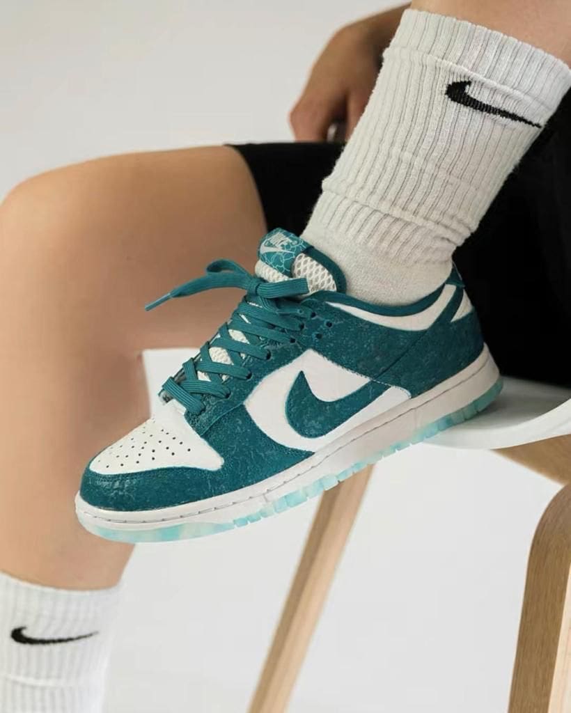 63f73022-9705-4c5d-8a77-2ccb9e68f2e2_e7e6b792-fd7f-47a0-865a-19bd5985b959 | AE JERSEY Teal and white Nike sneaker with white swoosh, worn with ribbed white socks, resting on a chair leg.