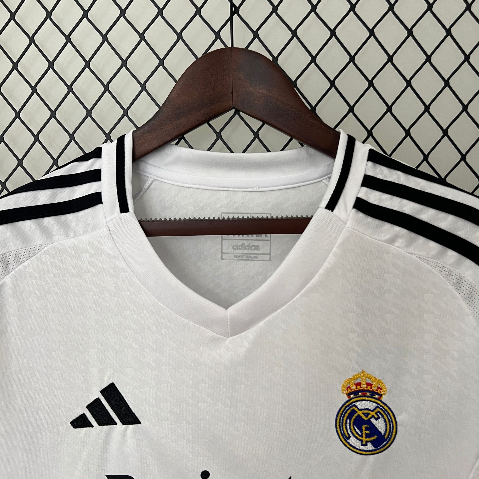 652fbda4 | AE JERSEY White Real Madrid soccer jersey on a wooden hanger, with the adidas logo and Real Madrid crest visible, set against a diamond-pattern chain-link fence.
