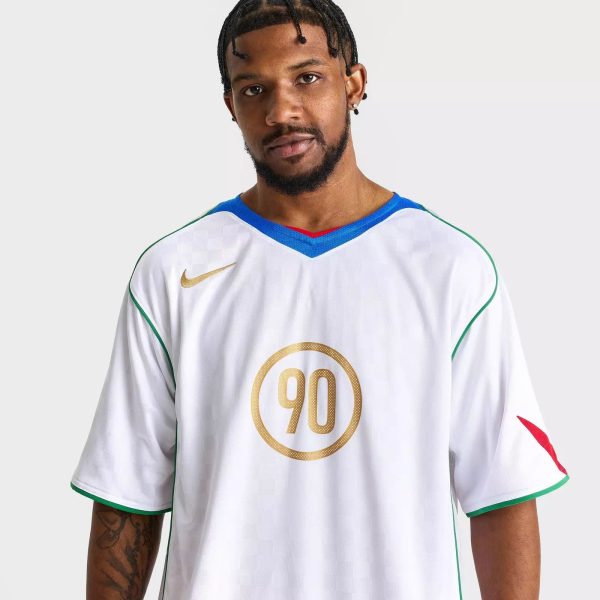 Portrait of a man with short dreadlocks and a beard, wearing a white sports jersey with blue trim and green piping, featuring a large gold circular 90 emblem on the chest against a light gray background.