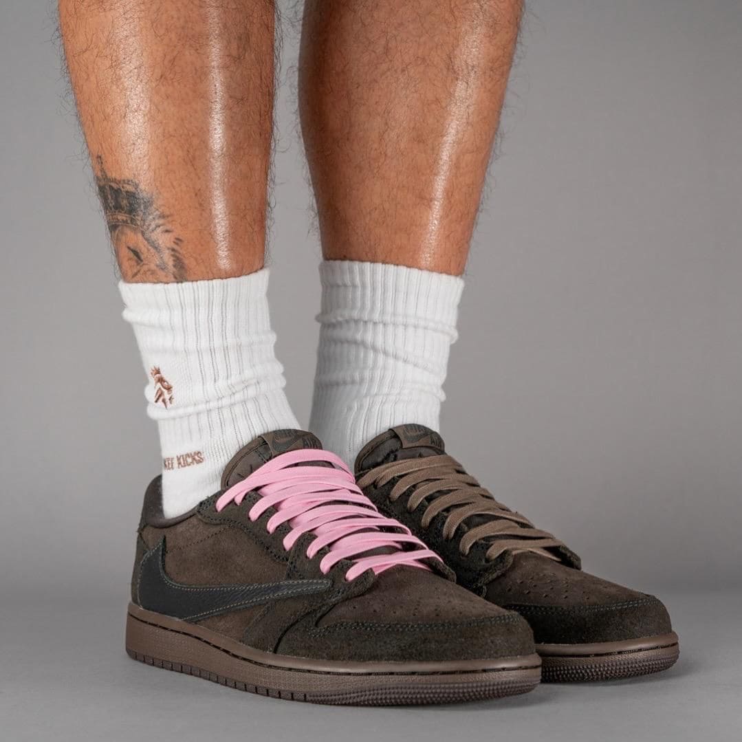 68e3711c-b248-4ee6-b618-aa19b6f4fc96 | AE JERSEY Brown suede sneakers with pink lace on the left shoe and brown lace on the right, worn with white crew socks and visible leg tattoos against a gray backdrop