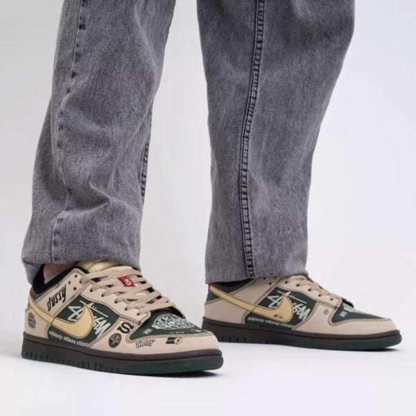 Low-top sneakers in beige and dark green with graffiti logos and a gold swoosh, paired with gray denim jeans.