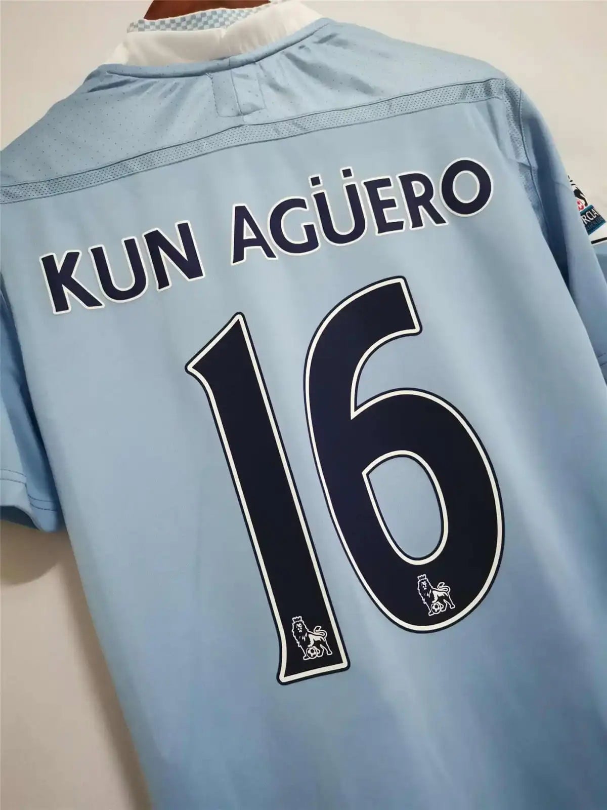 6b885e57-1 | AE JERSEY Back of a light blue soccer jersey with the name 'KUN AGÜERO' and the number 16, with small club crests visible near the bottom of the digits.