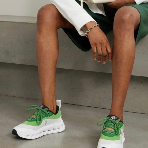 Person seated on a concrete surface, wearing a white blazer and green outfit, showcasing green-and-white athletic sneakers and holding a white bag.