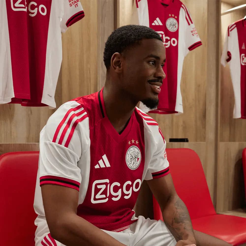 6e0097c8-b1e2-167c-a485-16725c3f96c3 | AE JERSEY Smiling man in a red and white Ajax jersey sits on a red bench in a locker room, with hanging jerseys behind him.