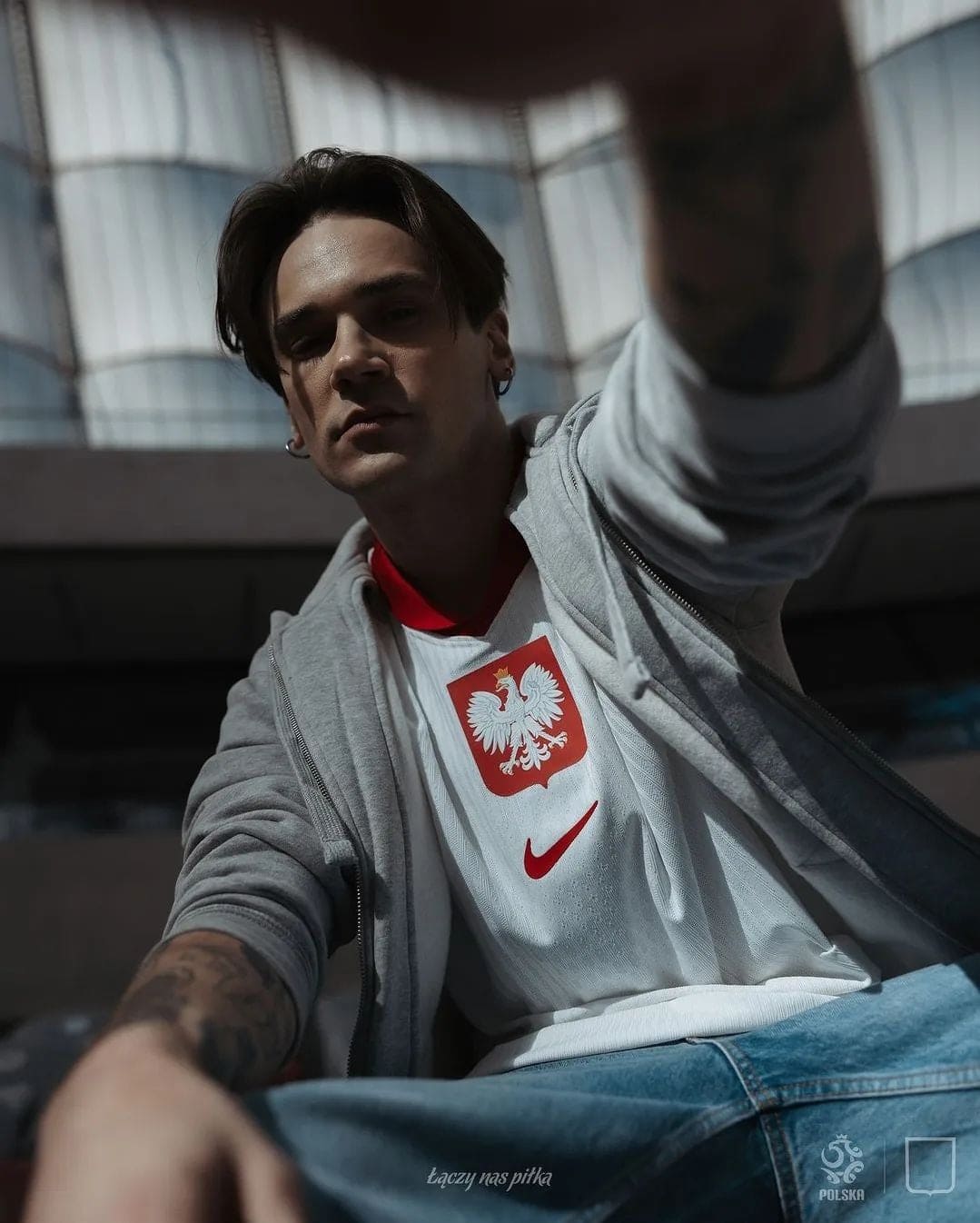 6hP4rzleBV9EUIV | AE JERSEY Young man wearing a white Poland football jersey with eagle crest, gray hoodie, taking a low-angle selfie outdoors.