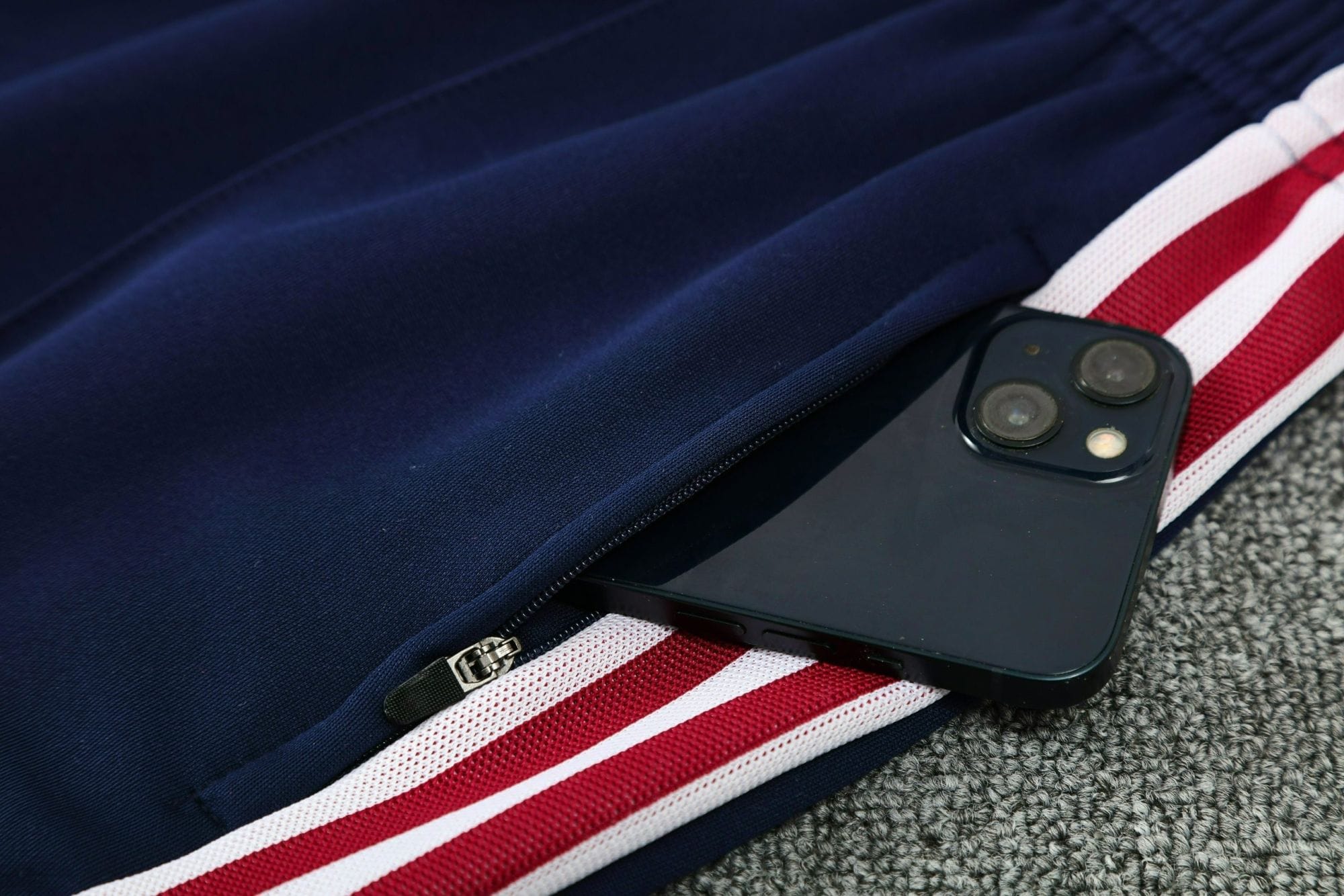 798e6097 | AE JERSEY Close-up of a navy backpack zipper with a black smartphone showing dual cameras resting on a red-and-white striped strap.
