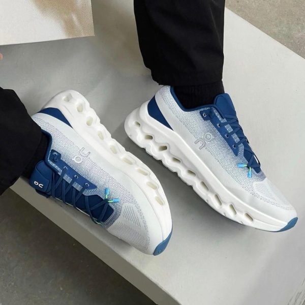 White and blue On running sneakers with a segmented hollow midsole, worn by a person in black pants.