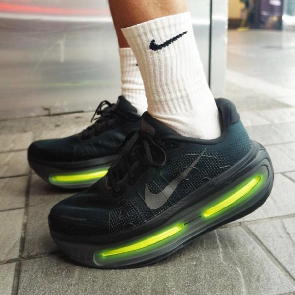 Black Nike running shoes with bright green translucent air sole, white crew socks, standing on a paved sidewalk.