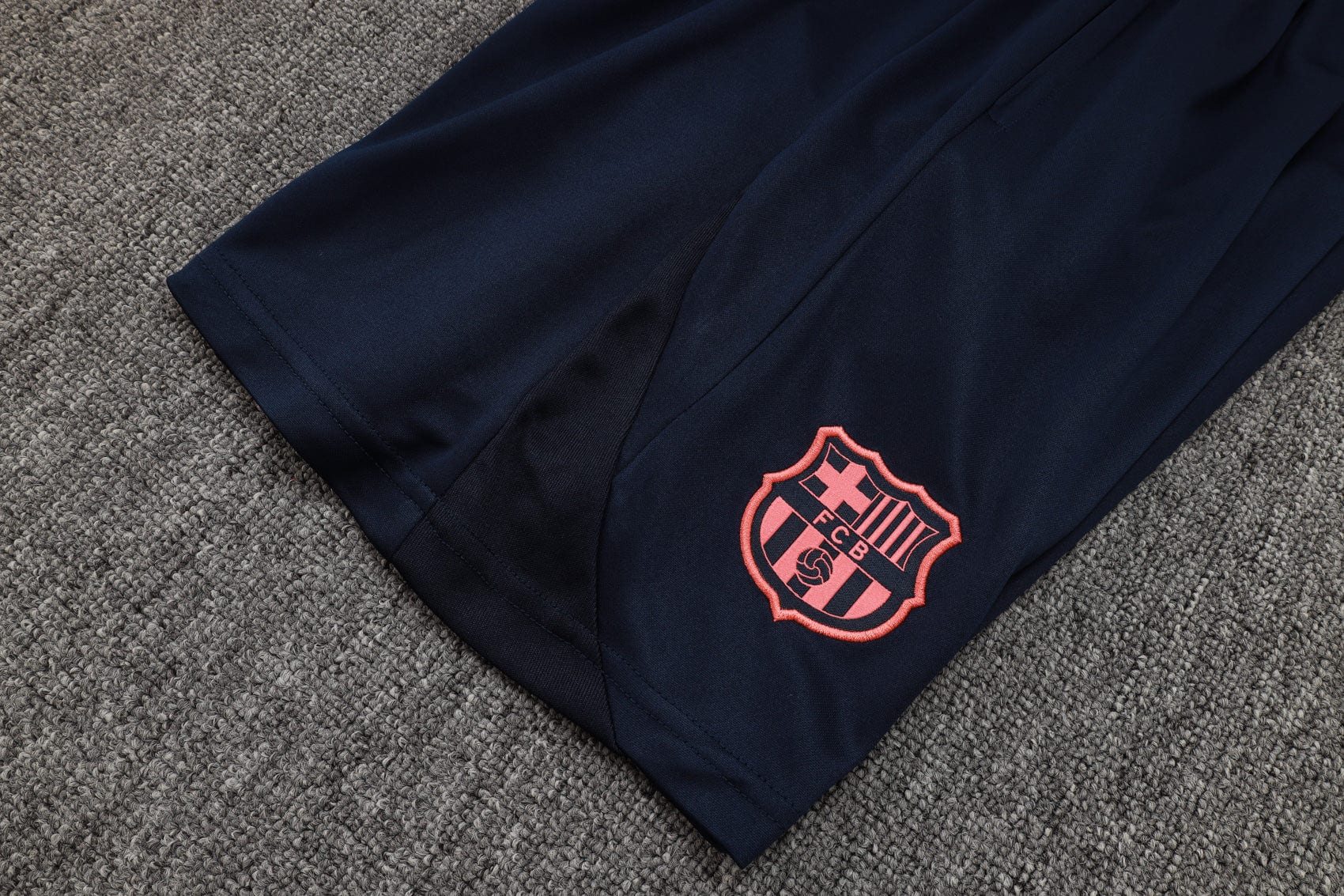 7d763b27 | AE JERSEY Navy athletic pants with a pink FC Barcelona crest embroidered on the lower leg, laid on a grey carpet.