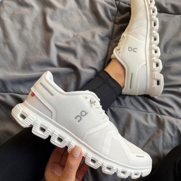 White On running shoes with a chunky segmented sole, one shoe resting on a bed and the other held in hand (casual indoor setting).