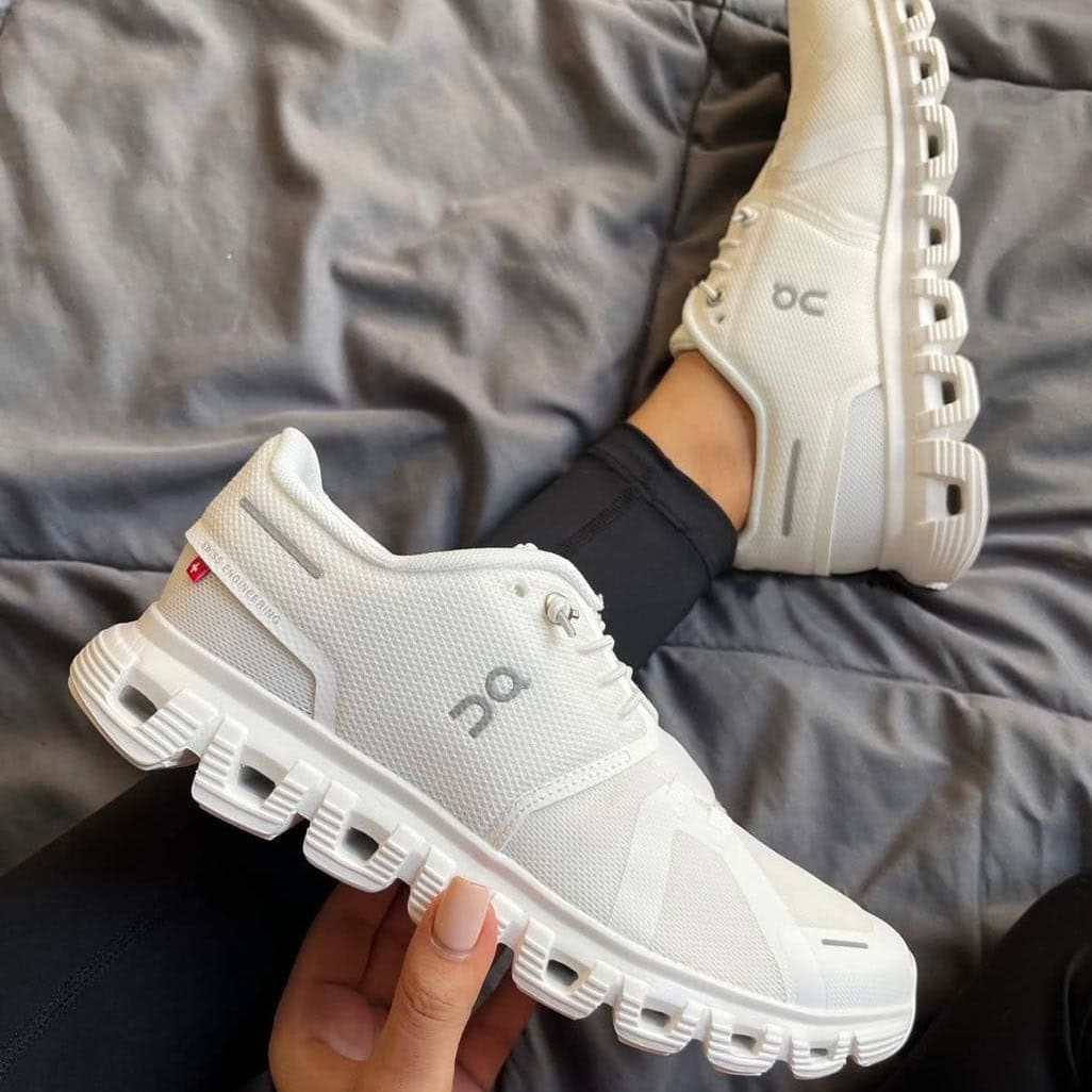 7ed704b3-0e80-4e9a-b14b-f3b1fc722cf7 | AE JERSEY White On running shoes with a chunky segmented sole, one shoe resting on a bed and the other held in hand (casual indoor setting).