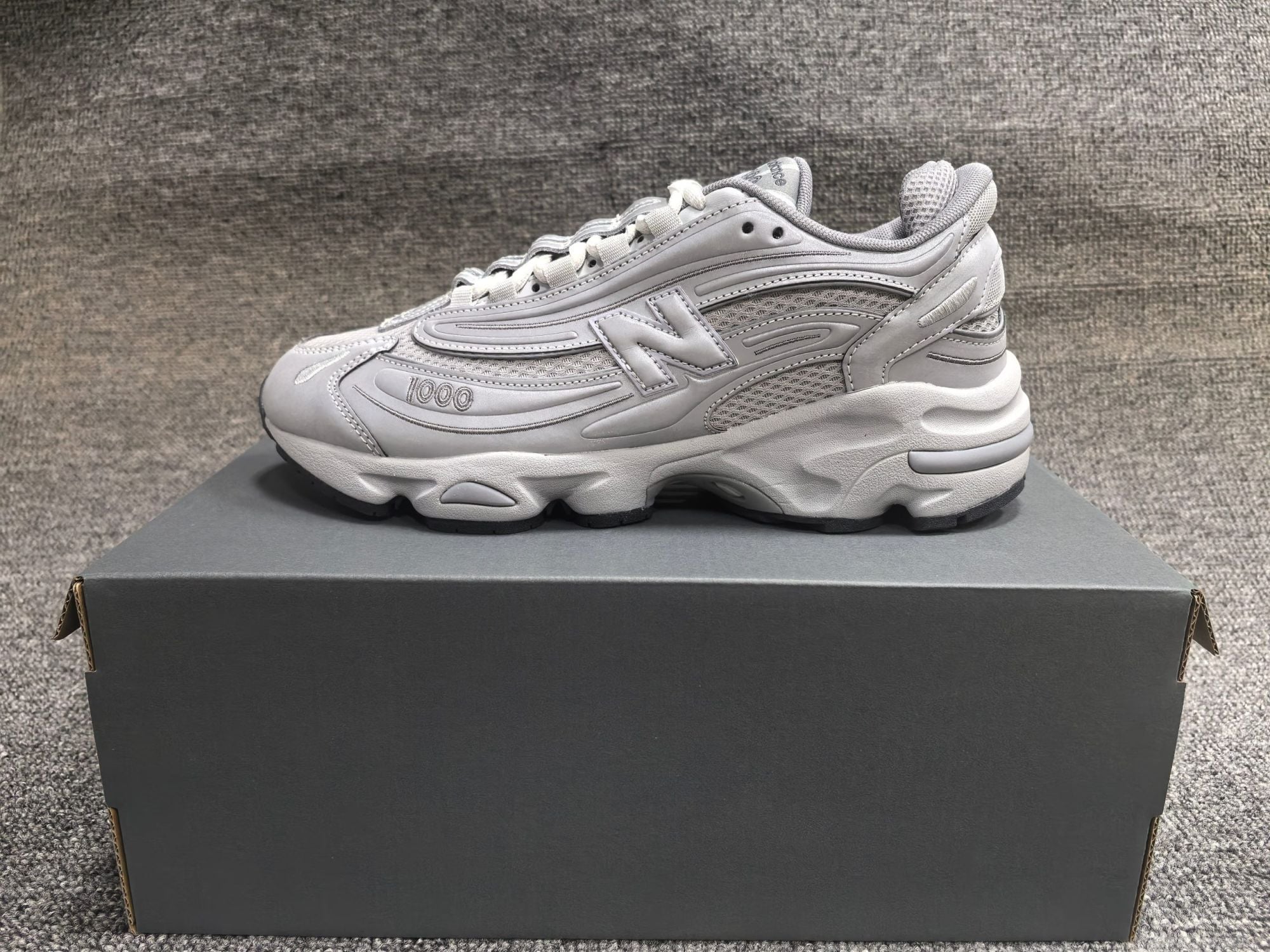 7f055ab0 | AE JERSEY Gray athletic sneaker shown in profile resting on a dark shoe box against a textured gray carpet.