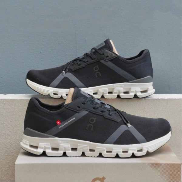 Pair of dark gray/black running shoes with thick white soles stacked on a box, showing the On logo and Swiss engineering tag.