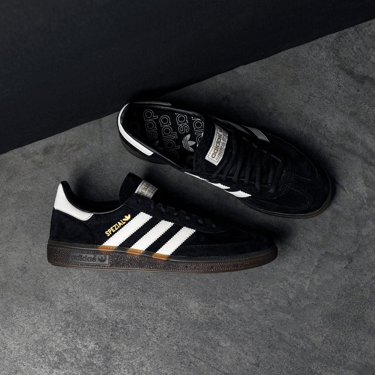 Black Adidas Spezial sneakers with white three stripes and brown sole on a gray surface, top view showing logo details for product photography