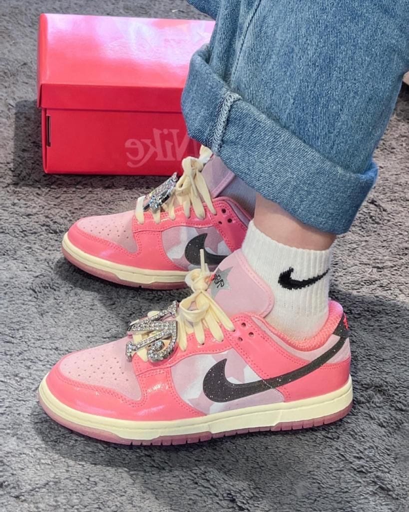 822a7860-5941-436a-bfcc-8b1f05726901_bcd2de48-06f5-4353-ab66-d8259439db3e | AE JERSEY Pair of pink Nike sneakers with white laces and rhinestone embellishments, worn with rolled-up jeans and white Nike socks on a gray carpeted floor, pink Nike box in the background