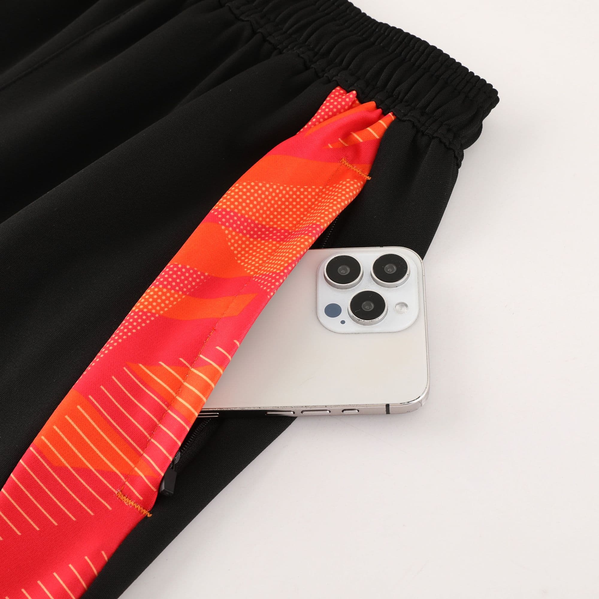 833228e8 | AE JERSEY Close-up of a black fabric with an elastic cuff and a bright orange patterned strap lying across it, next to a silver smartphone with three cameras.