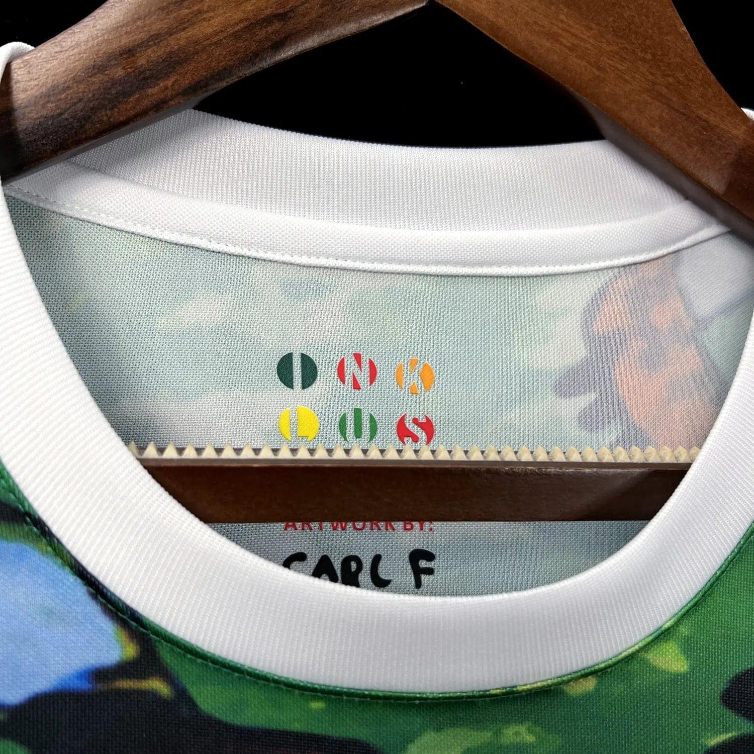87b93b94933f34c0 | AE JERSEY Close-up of a shirt collar on a wooden hanger, revealing a multicolor 'ONE OF A KIND' design printed on the inside neck label.