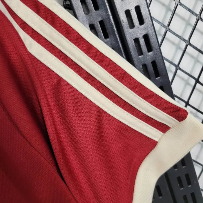 8f3b906cd67f0741_2ddfffba-51fb-4fe4-abe6-48fa36fba390 | AE JERSEY Close-up of a red athletic jersey sleeve with white double piping on a cream trim, hanging on a metal rack.