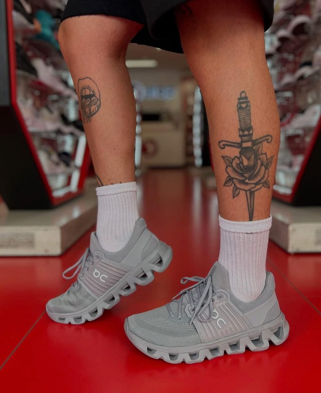 93b251e2-e8e3-4c60-b22b-fe209c4253a9 | AE JERSEY Person standing in a store wearing gray athletic sneakers and white socks; left calf has a circular tattoo, right calf has a dagger and rose tattoo.