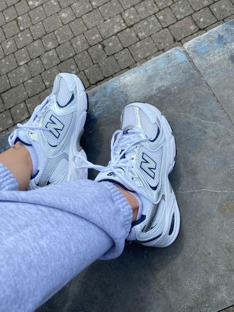 941c4b8d-1345-4ebb-b0ae-1248268cdd1b | AE JERSEY Person wearing white New Balance sneakers and light gray sweatpants, standing on a paved sidewalk.