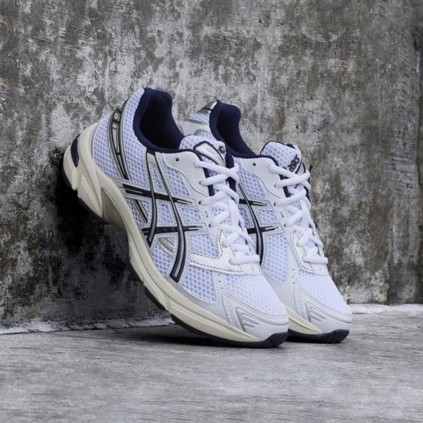 Pair of white and navy ASICS running shoes with mesh panels leaning against a textured concrete wall.