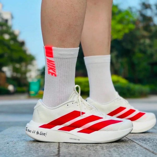 993be5cb-ff05-4a2c-8f70-5c4c8a78da23 | AE JERSEY Person standing outdoors in white running shoes with red stripes and white socks; the shoe sole reads “To run is to Live!”