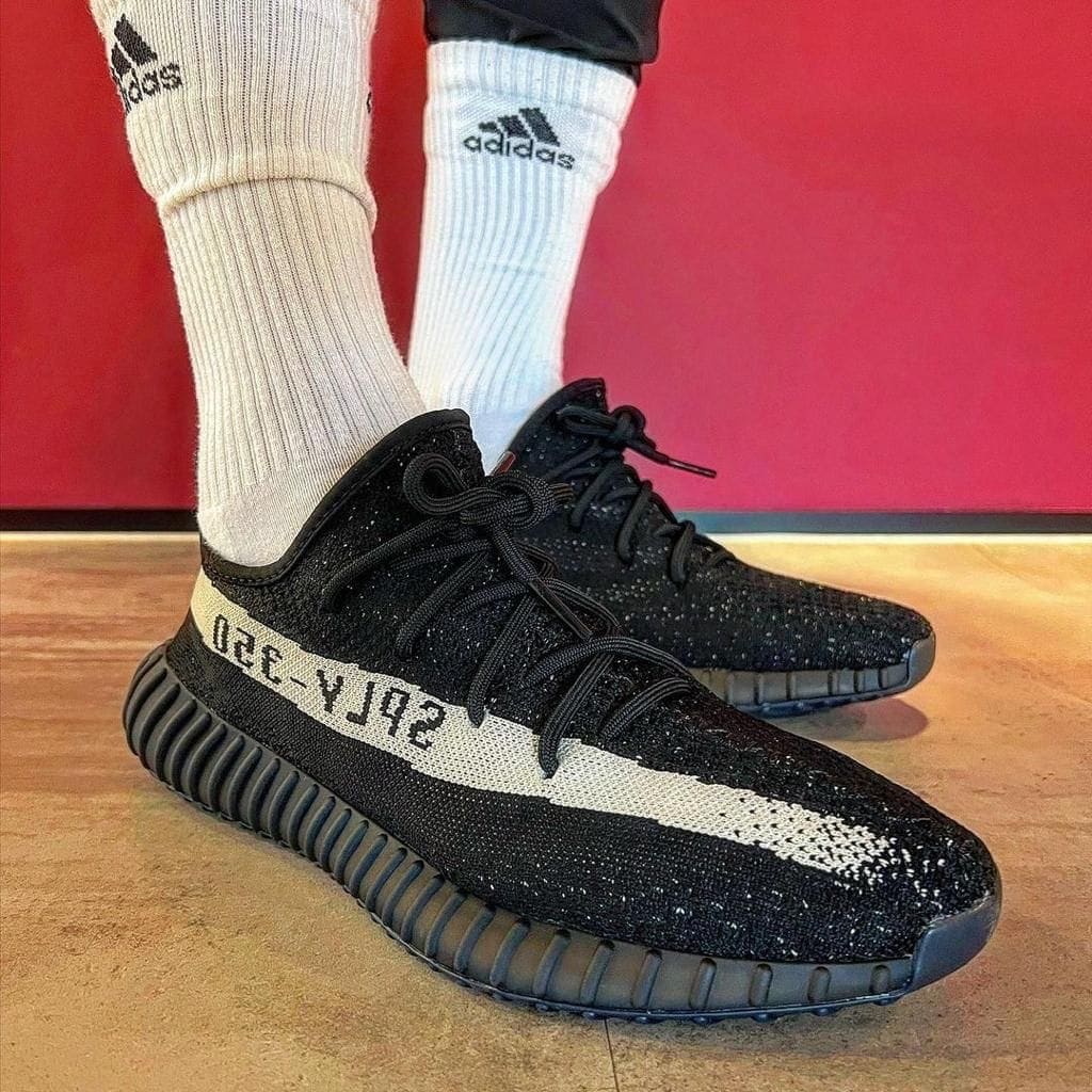 998c91e0-d727-4335-8bea-1099bab342a2_a0227e0b-7721-4333-8bfa-0b05778b35b0 | AE JERSEY Black knit sneakers with a white SPLY-350 stripe, worn with white Adidas socks on a wooden floor.